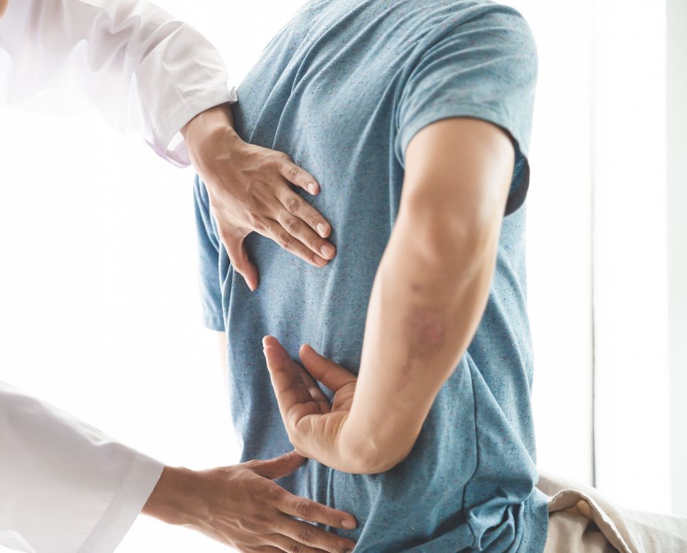 Doctor examining a person's lower back, possibly for pain. The person wears a blue shirt. The doctor wears a white coat.