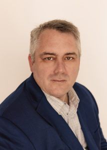 Man in a blue blazer and patterned shirt looks directly at the camera against a white background.