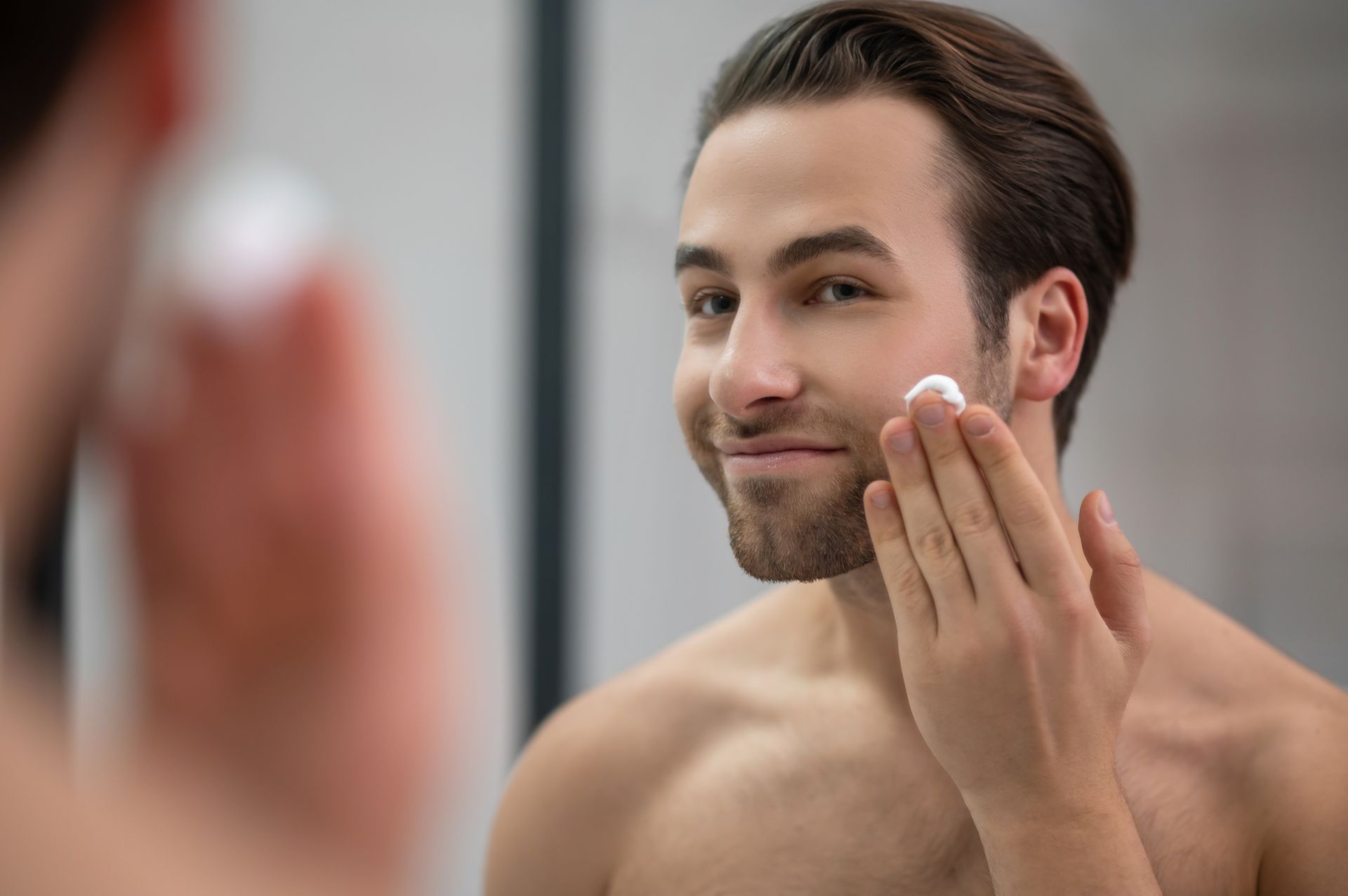 A shirtless man is applying lotion to his face in front of a mirror.