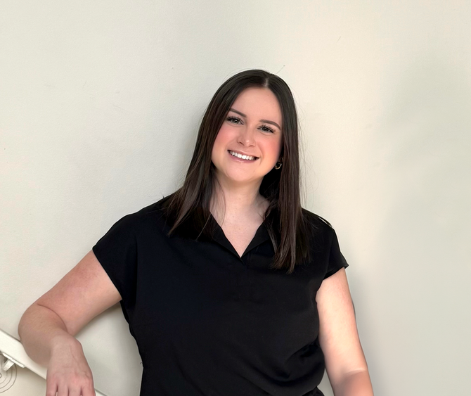 A smiling person with long brown hair, wearing a black shirt, leaning against a white railing against a plain wall.