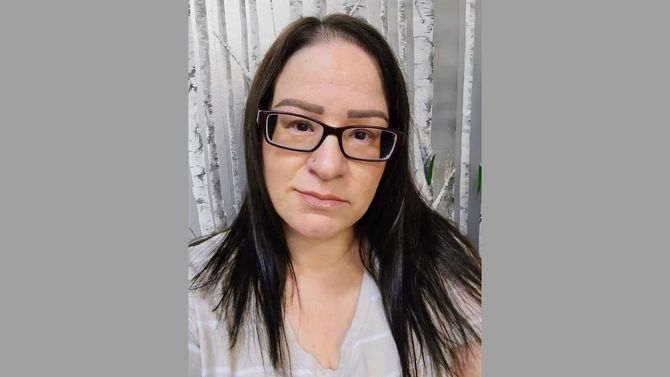 Person wearing glasses and a white top, standing in front of a light gray curtain.