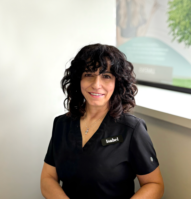 Woman with dark curly hair in black scrubs smiles near a window.