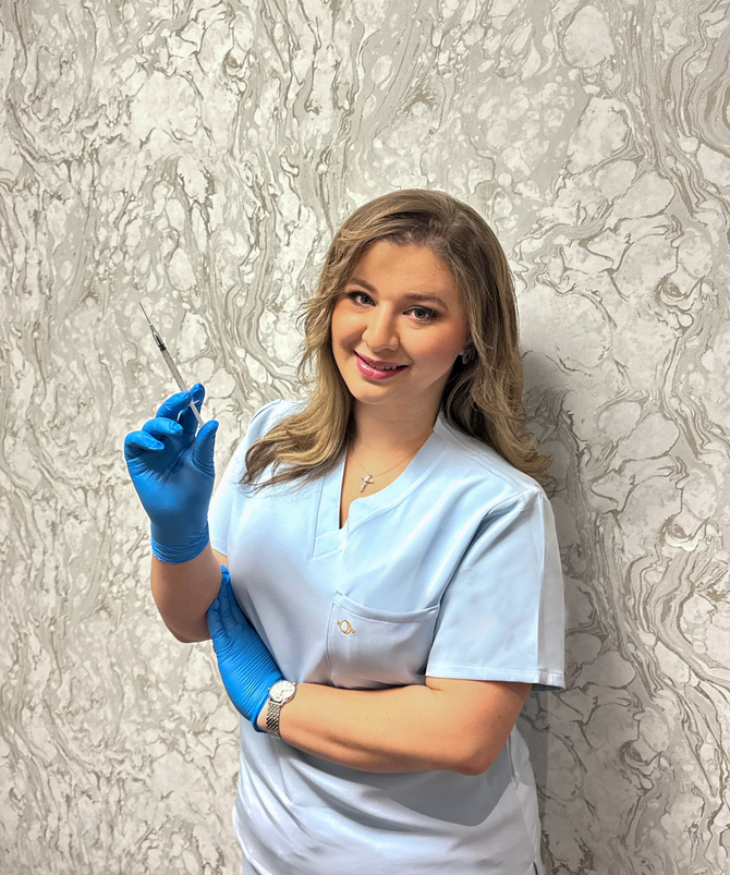 A healthcare professional in blue scrubs and gloves holds a syringe, smiling against a marbled patterned wall.