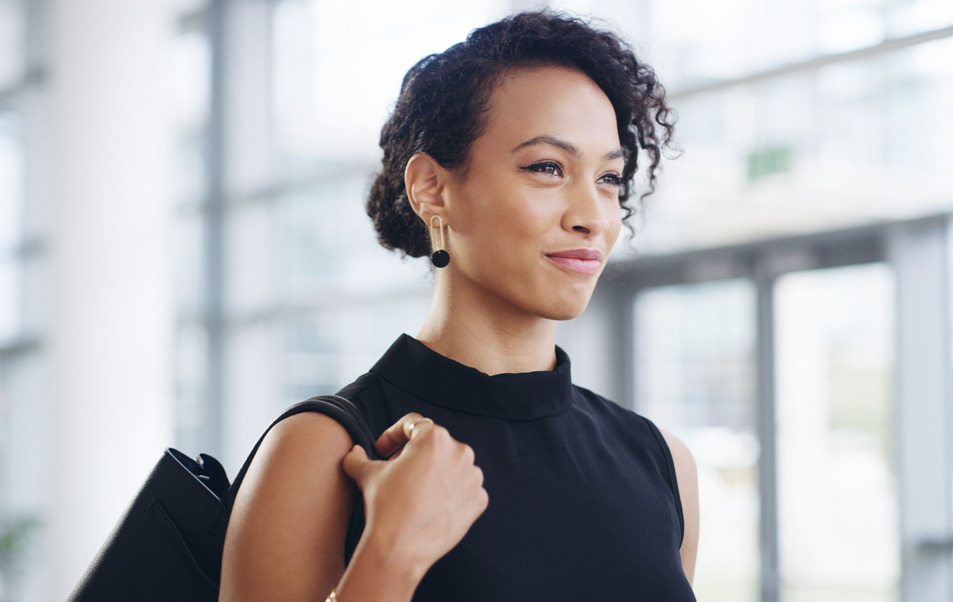 Woman in black dress and earrings smiles, carrying a bag in a modern office space.