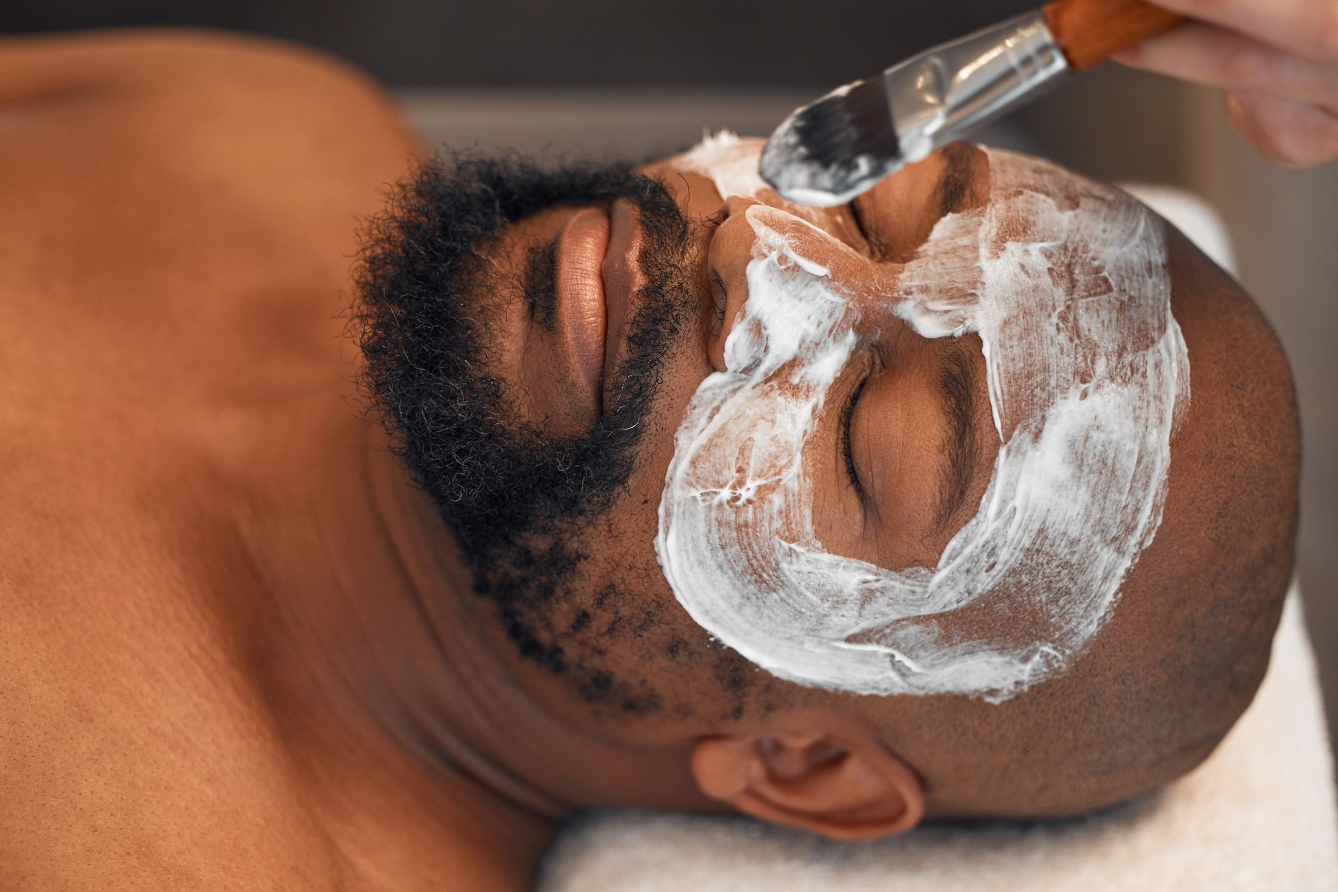 Man receiving a facial treatment with white cream applied and a brush on his forehead