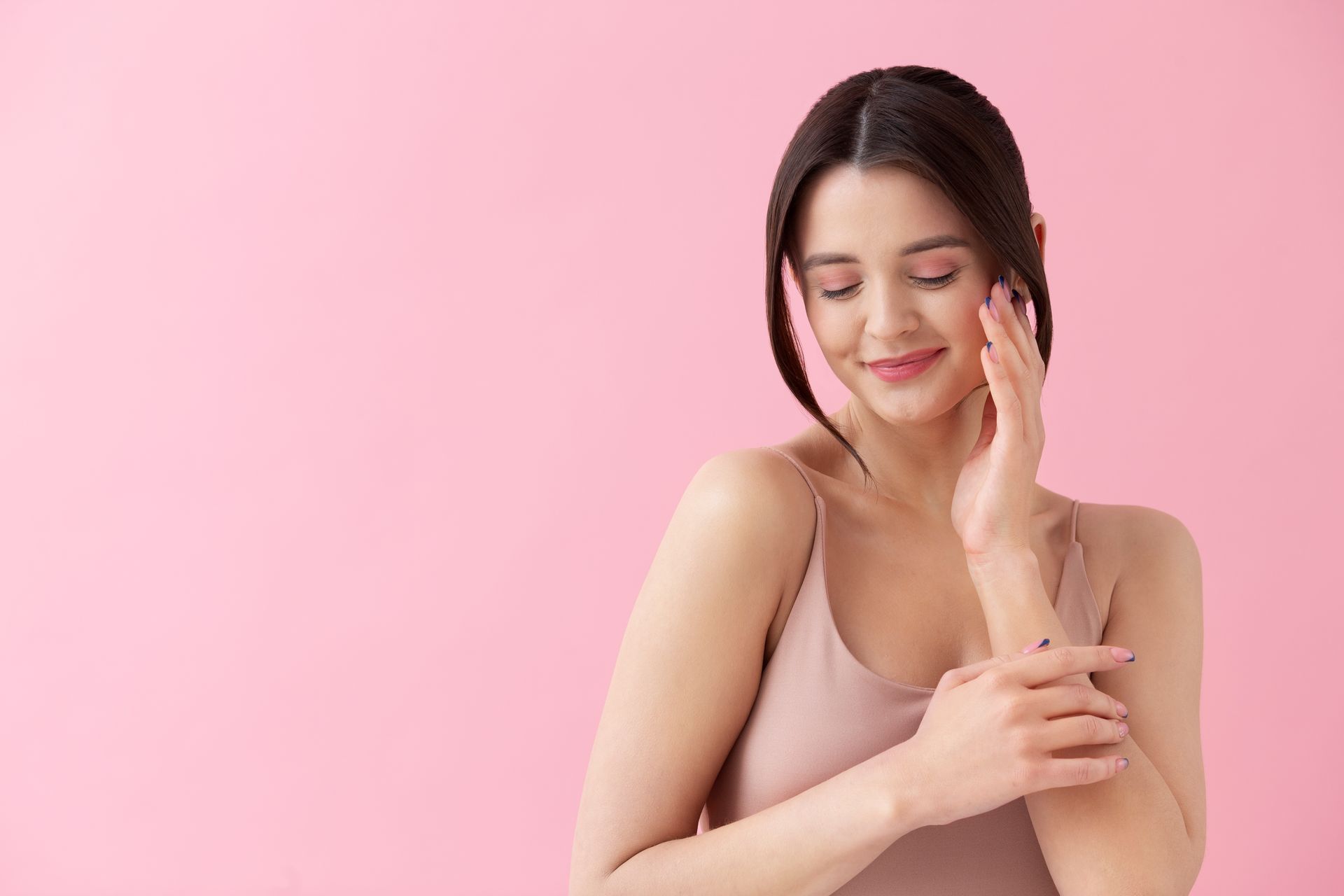 Woman in a pink tank top gently touching her face and arm against a pink background.