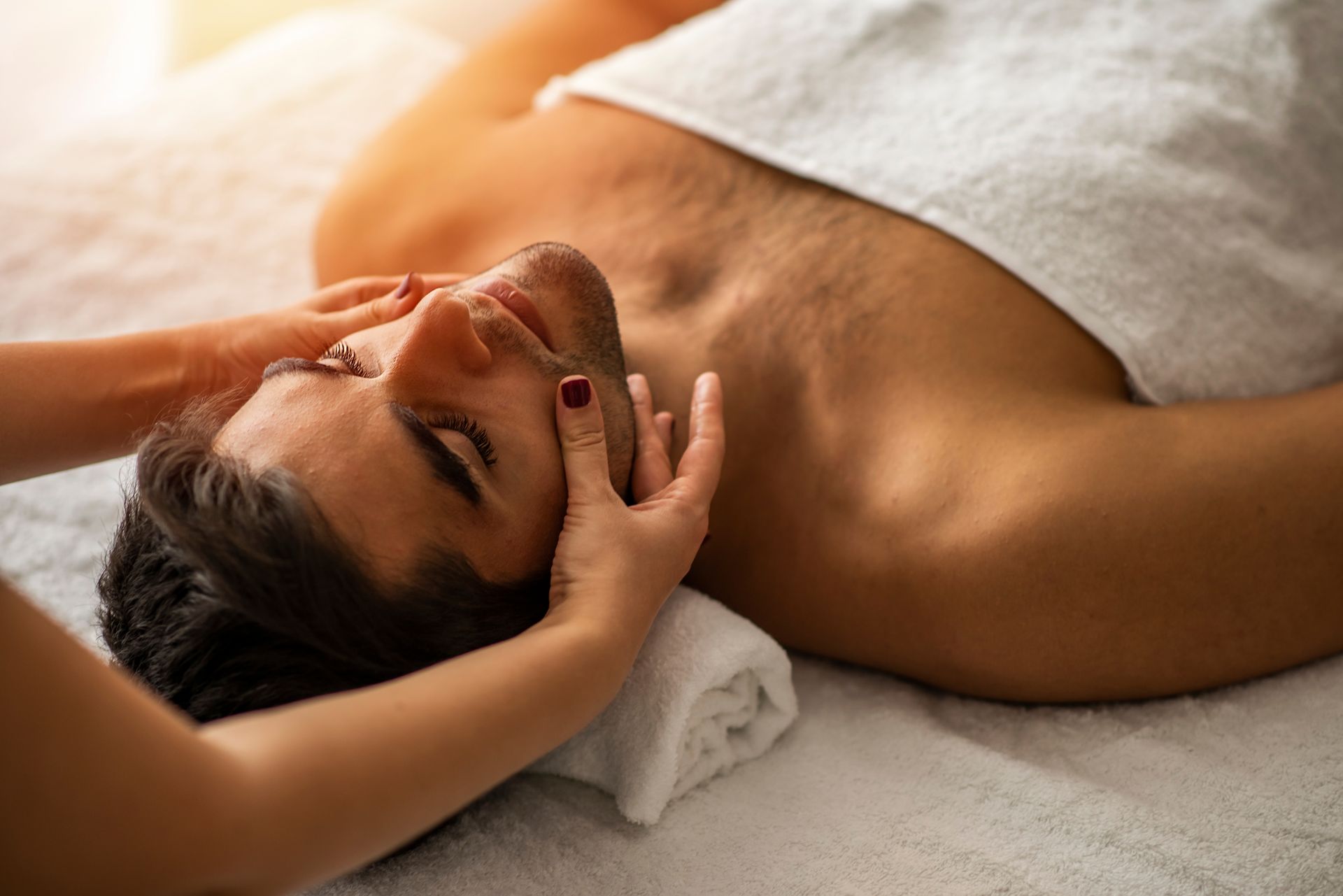 Man receiving a facial massage at spa; white towel, hands on face.