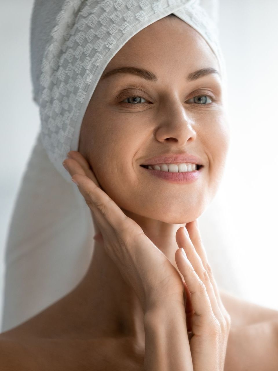 A woman with a towel wrapped around her head is smiling and touching her face.