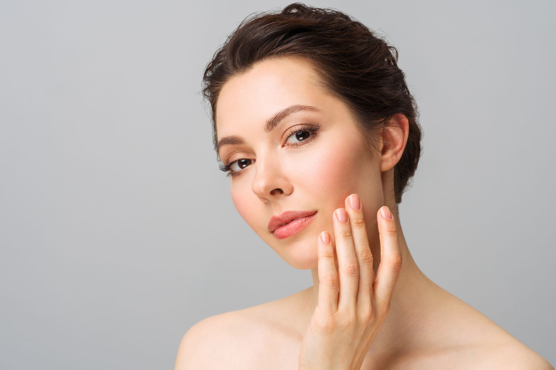 Woman touching her cheek, looking toward the camera. Neutral background, soft lighting.