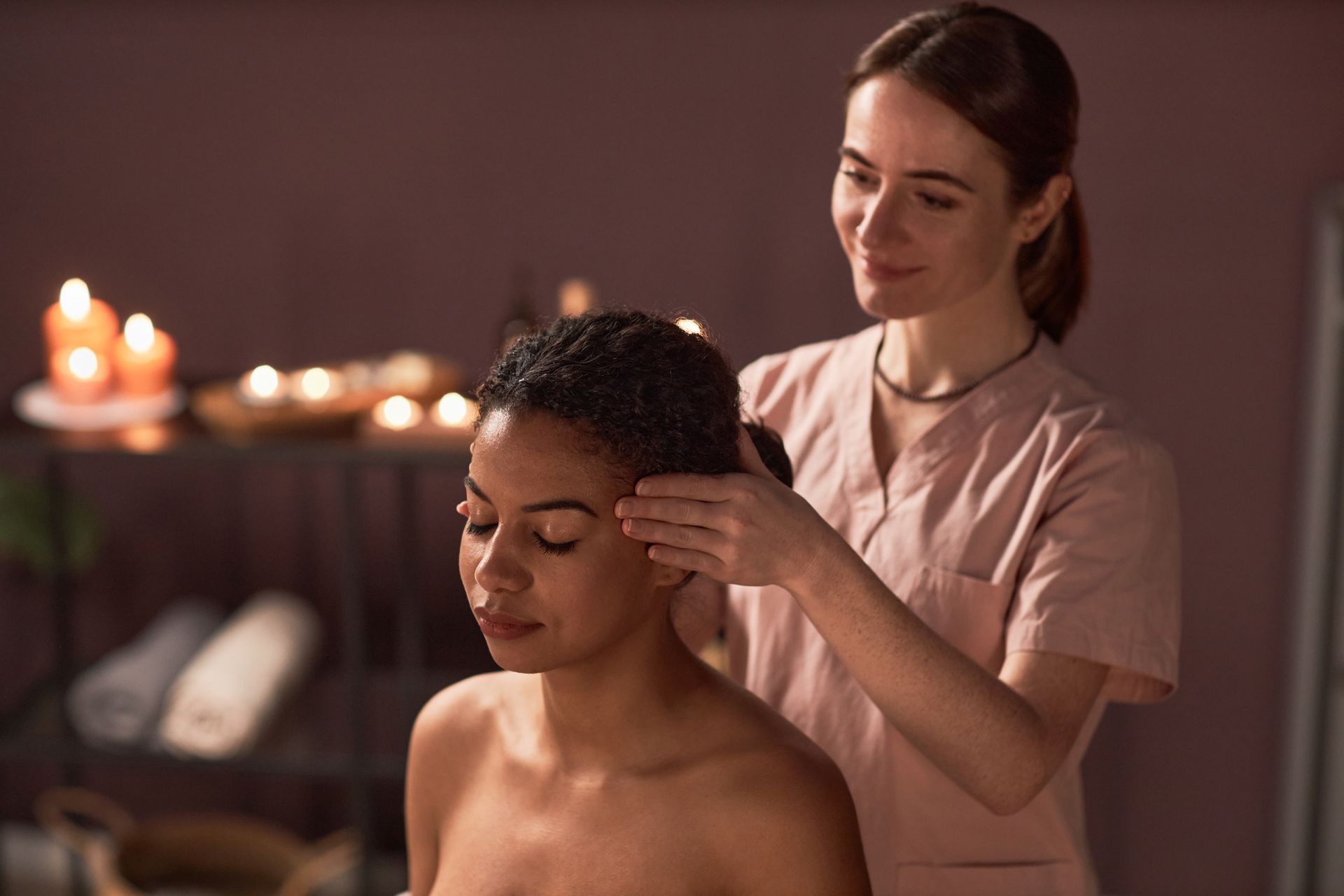 Woman receiving a head massage in a dimly lit spa. A masseuse is gently touching her hair.