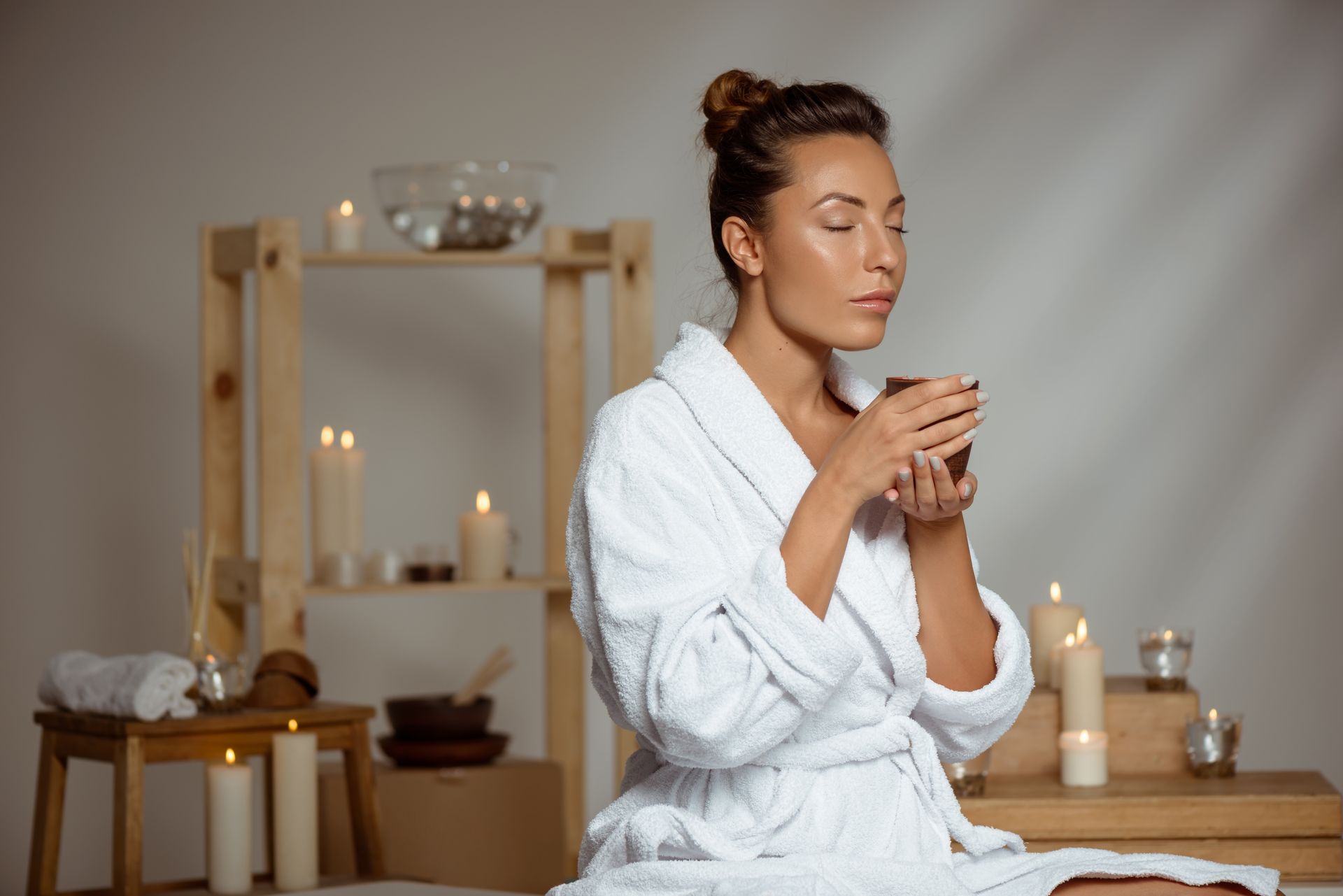 Woman in white robe sipping from a cup, eyes closed, surrounded by candles in a spa-like setting.