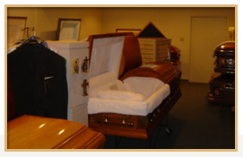 Funeral home display: open wooden casket with white interior, other caskets, suit, and framed art in room.