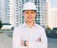Man in hard hat and white shirt, arms crossed, smiling in front of a building.