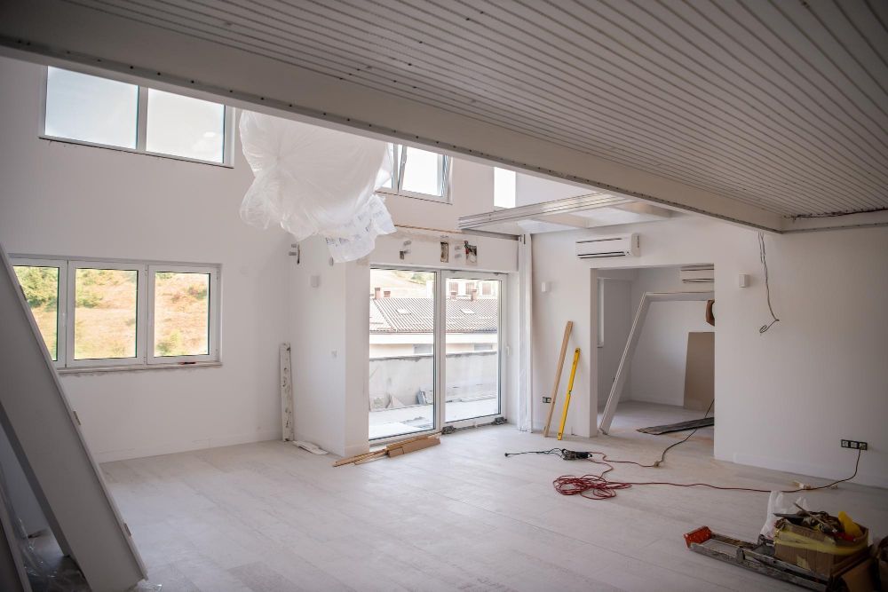 Interior view of a room under renovation with white walls, windows, and exposed ceiling, construction tools scattered.