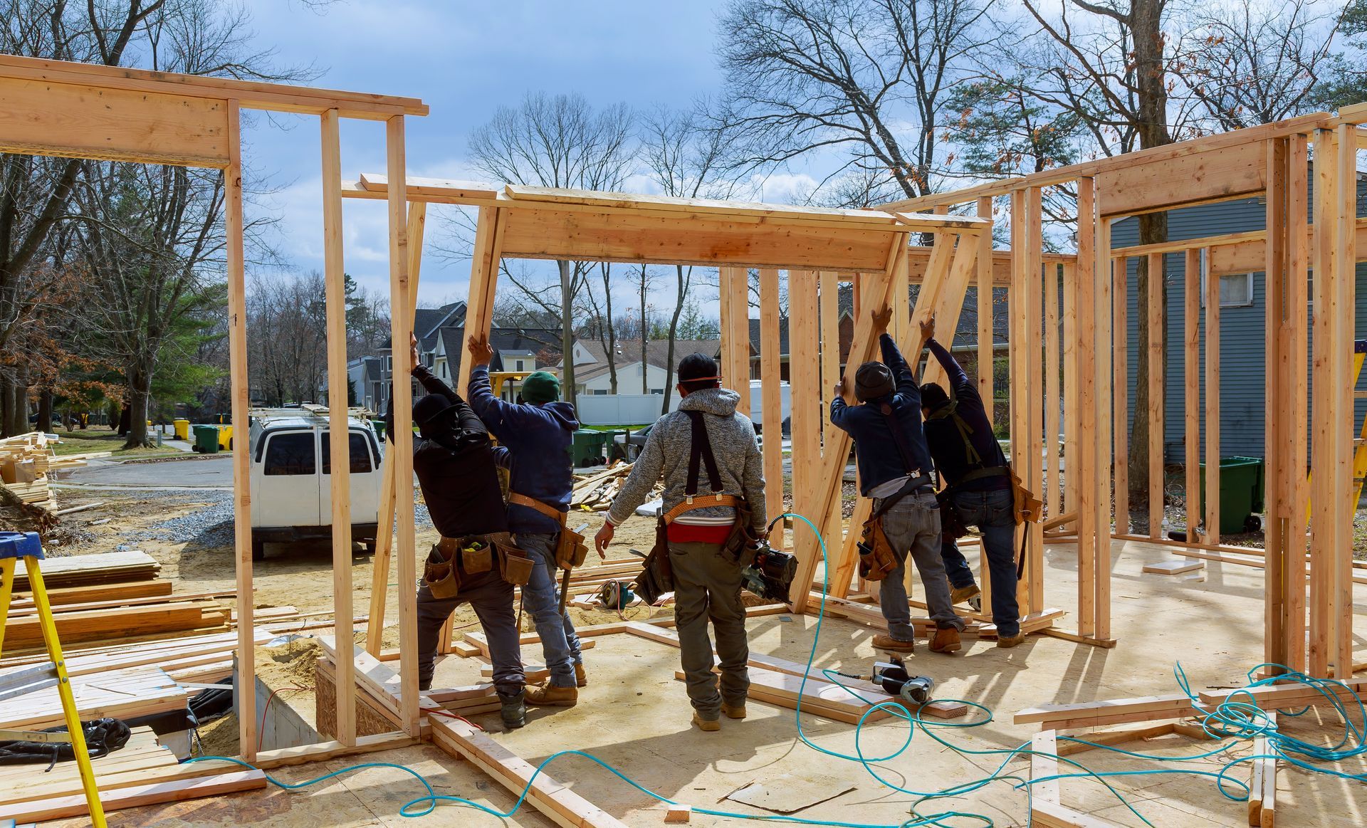 Construction workers raising a wooden wall frame at a building site; outdoors, sunny day.