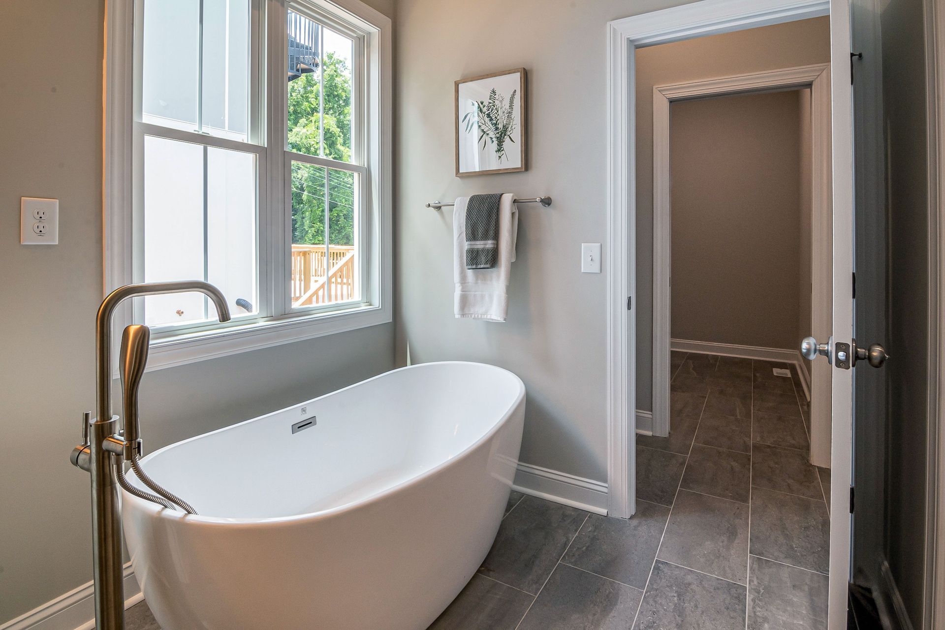 Modern bathroom with a white freestanding tub, brass faucet, window, and gray tile flooring