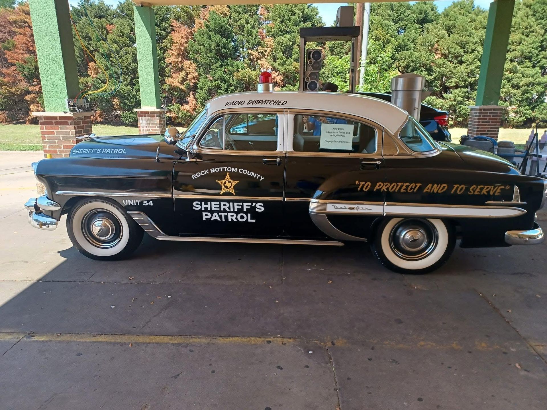 Classic black and white vintage police car with white-wall tires, parked under a covered area. Reads 