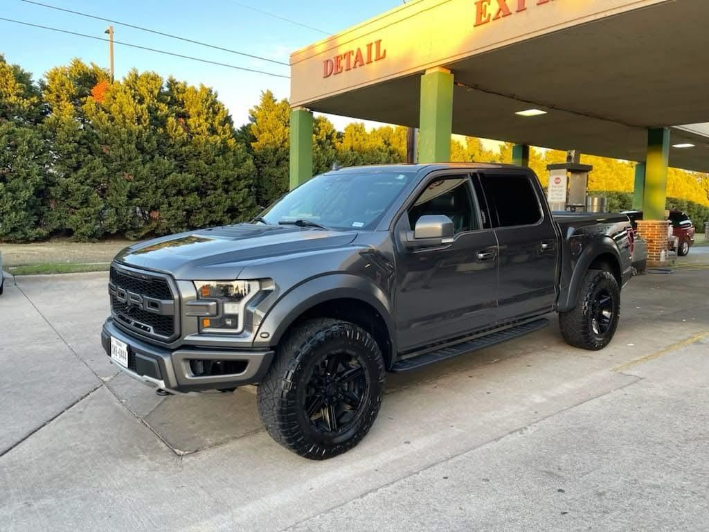 Gray Ford Raptor pickup truck parked under a car wash awning. Black wheels, daytime.