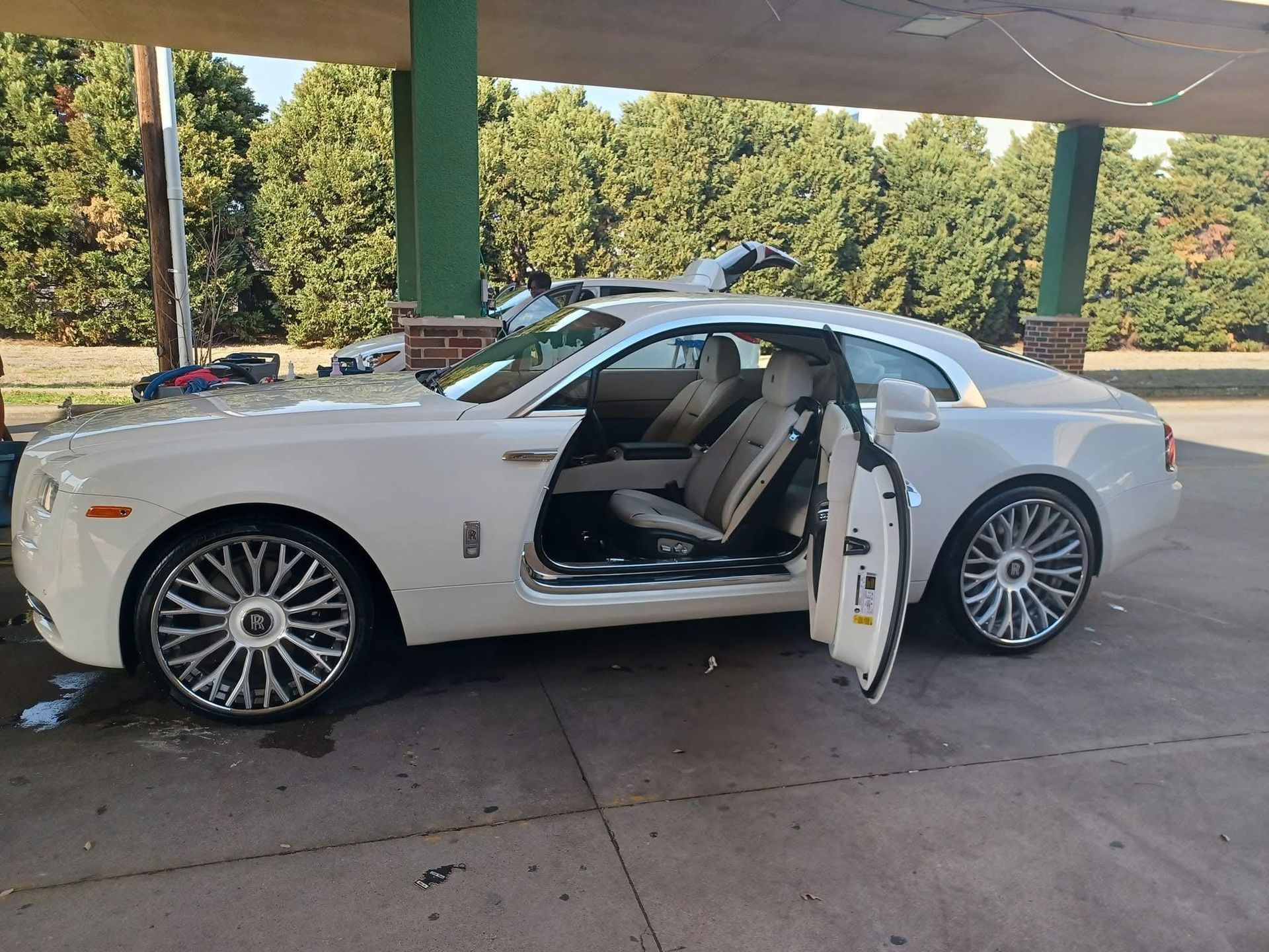 White Rolls-Royce with open doors at a car wash, reflecting a bright sunny day. The car has large chrome wheels and tan interior.