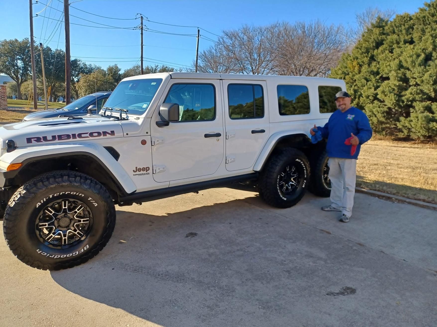 White Jeep Rubicon parked outdoors with a man in a blue shirt and cap standing next to it on a sunny day.