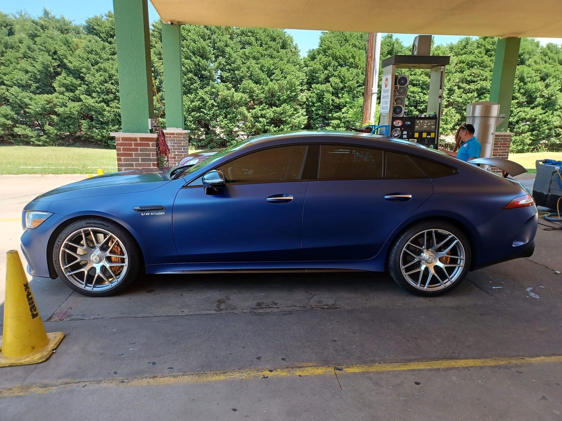 Blue Mercedes-AMG GT 4-door coupe at a gas station under a canopy.  It has chrome wheels and tinted windows.
