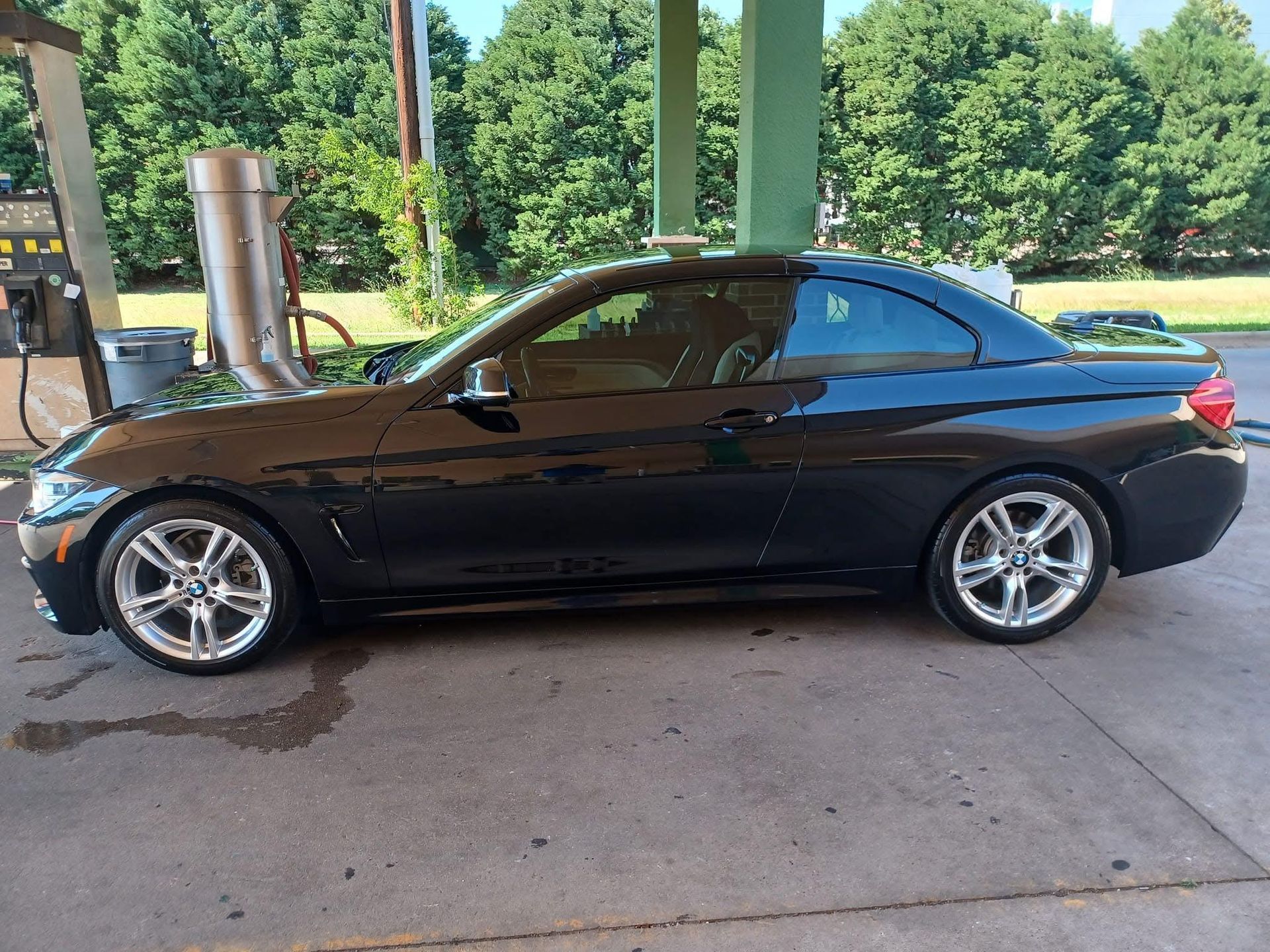 Black BMW coupe parked at a gas station, reflecting chrome wheels, with green trees in the background.