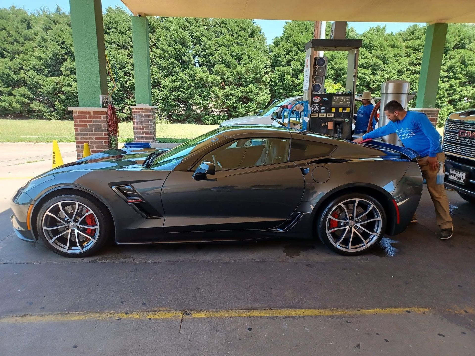 A gray sports car being washed at a gas station by a man in a blue shirt on a sunny day.