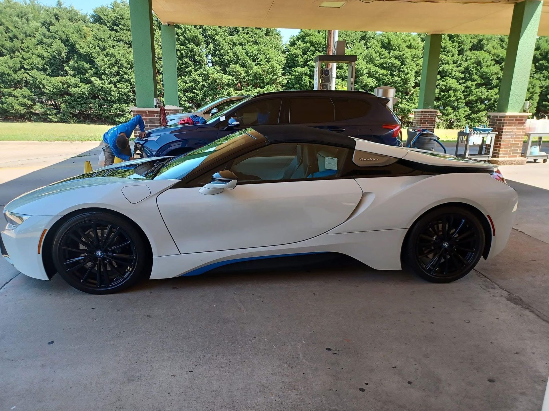 White BMW i8 convertible with black wheels parked under a canopy with a blue car and trees in the background.