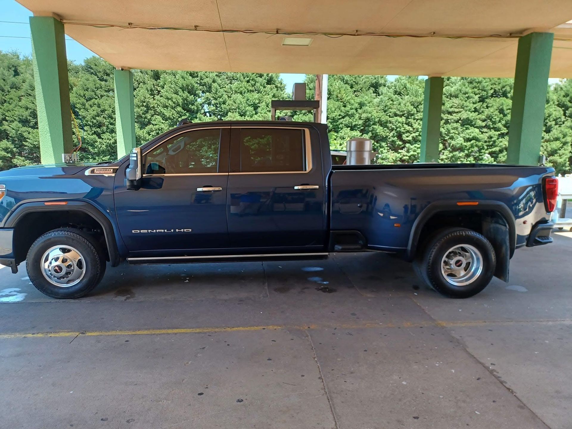 Dark blue Chevrolet Silverado 3500 dually truck parked under a covered structure, with trees visible in the background.