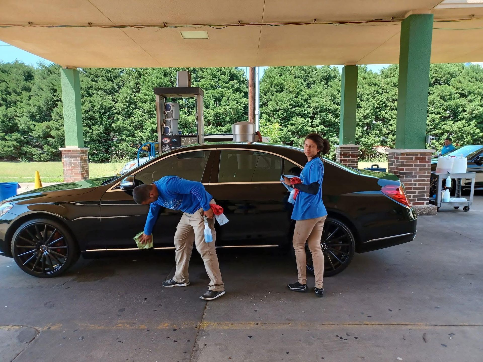 Two people washing a black car at a car wash. One person wipes the car's side, and the other holds cleaning supplies.