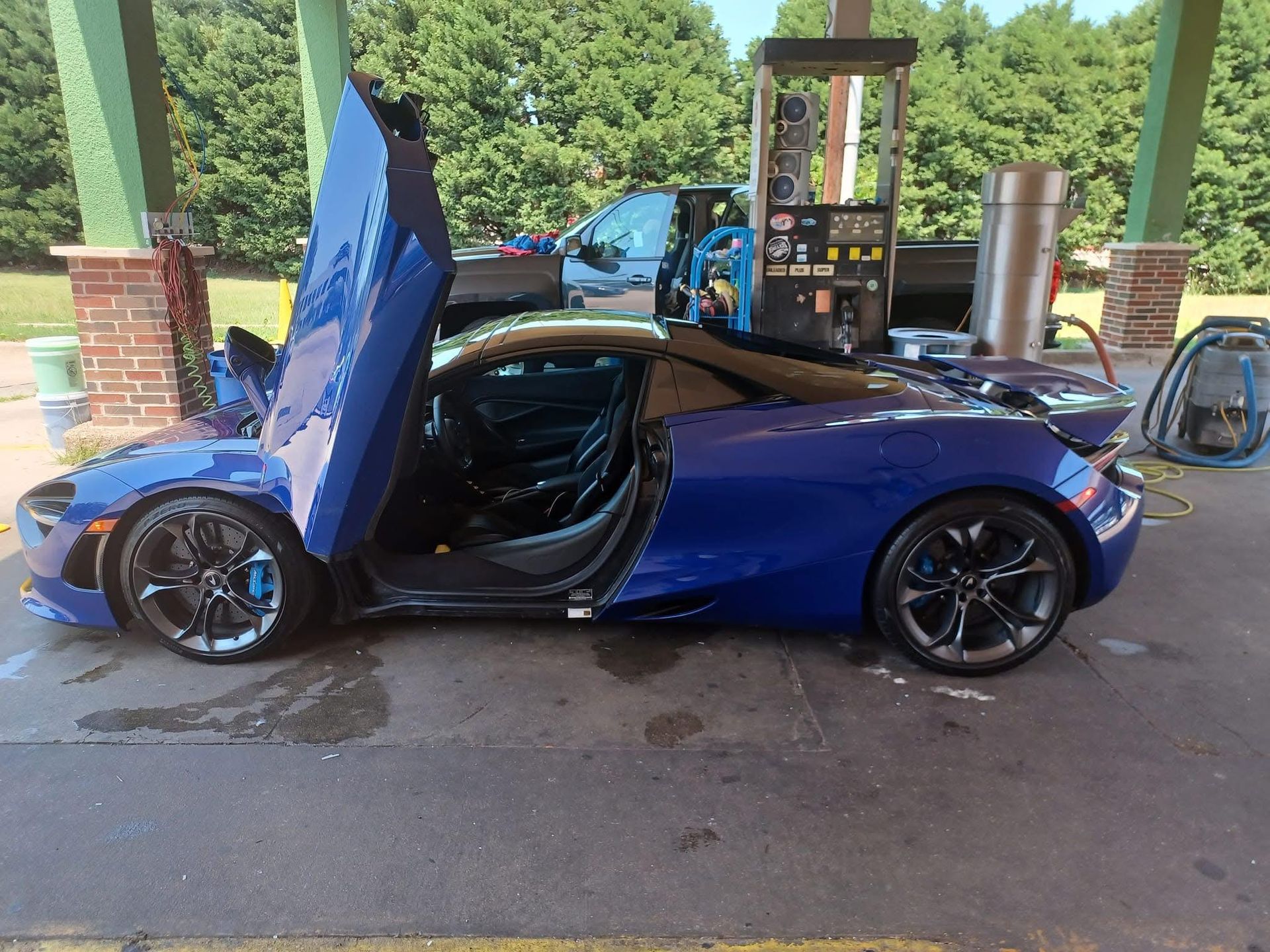 Blue McLaren sports car with doors open at a gas station. It's wet, and there's a dark roof and black wheels.