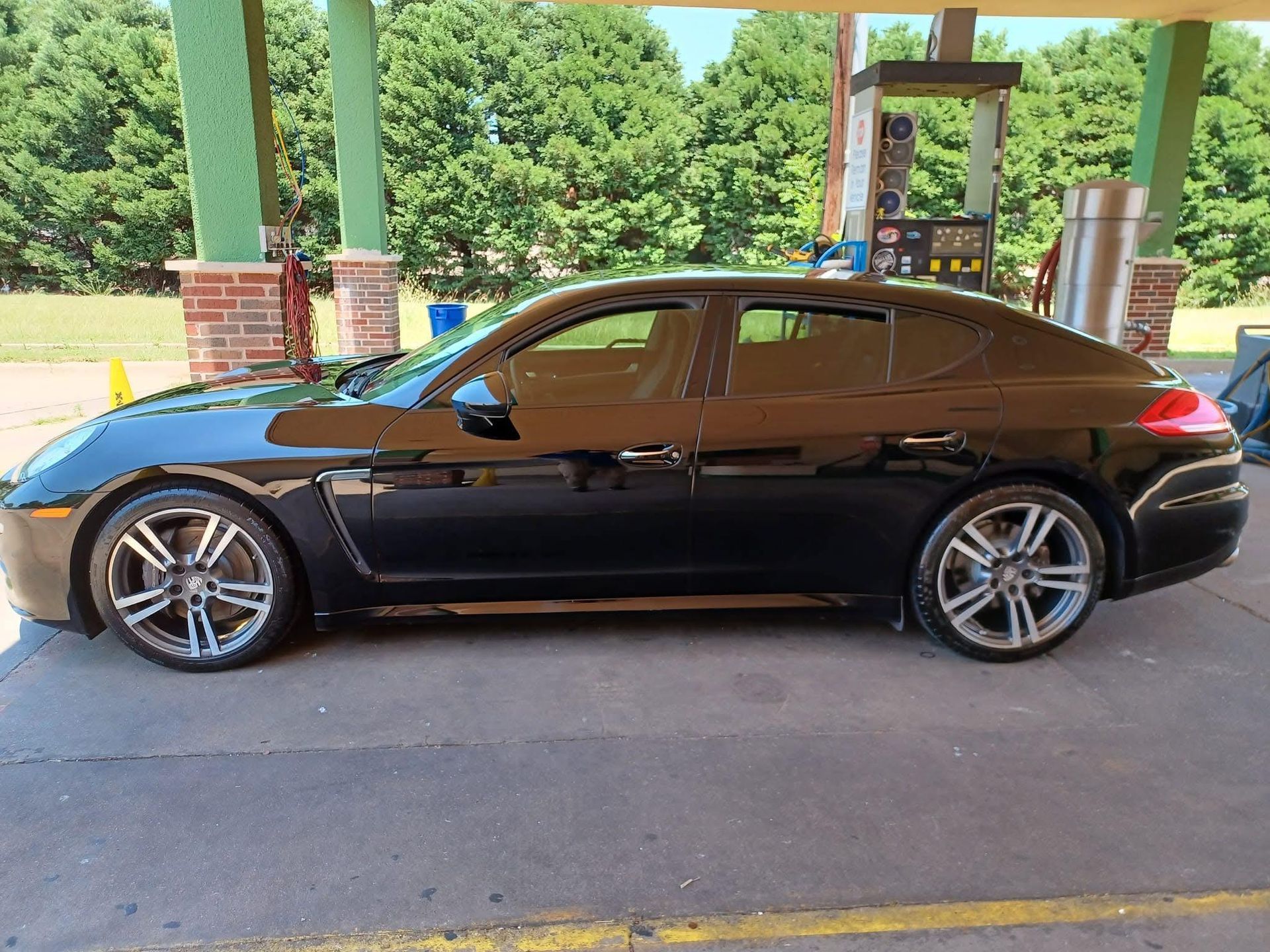 Black Porsche Panamera parked at a gas station. The car is in front of a gas pump under a green canopy.