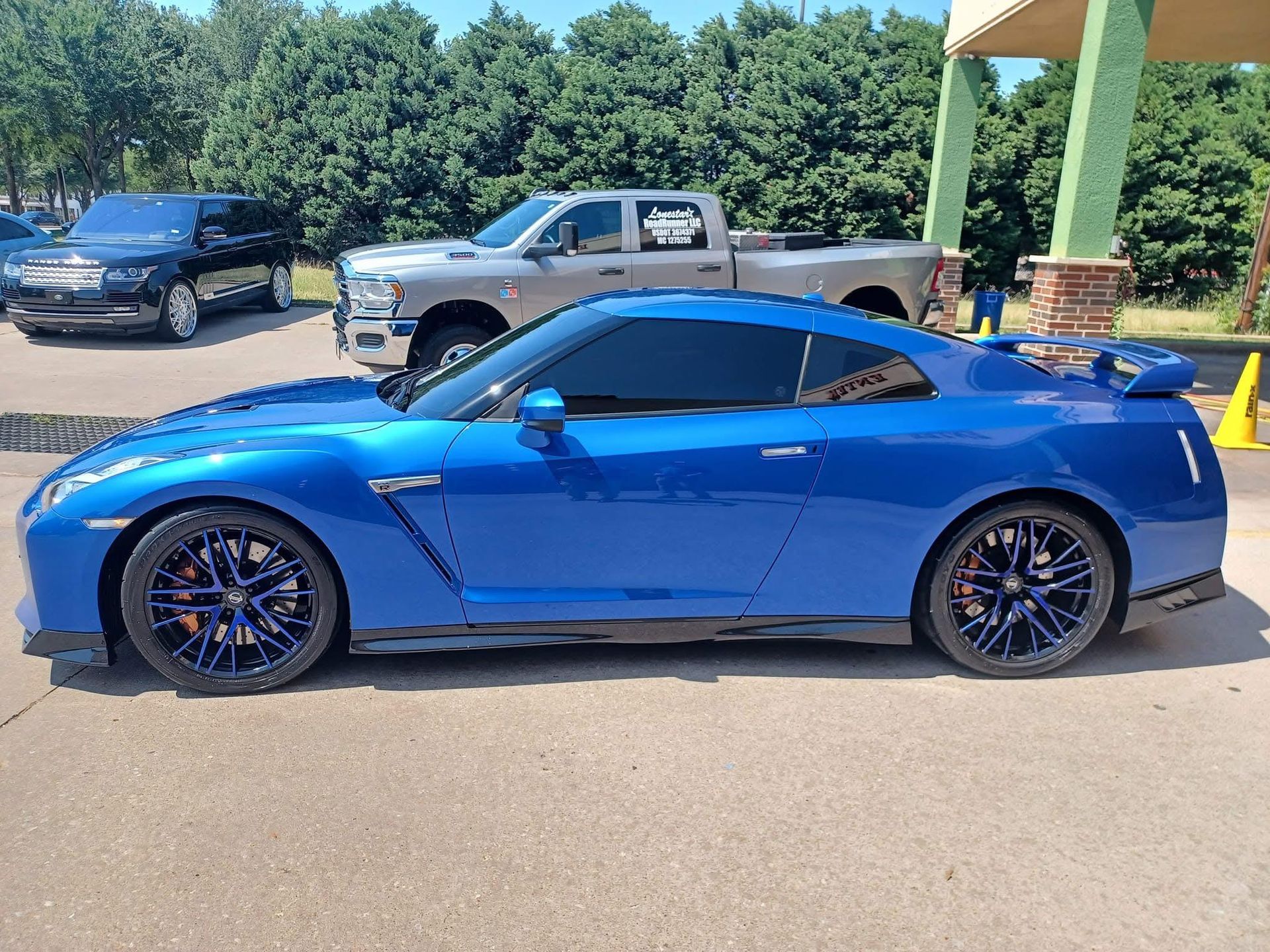 Blue sports car with tinted windows and dark blue rims parked outside. A truck and SUV are in the background.