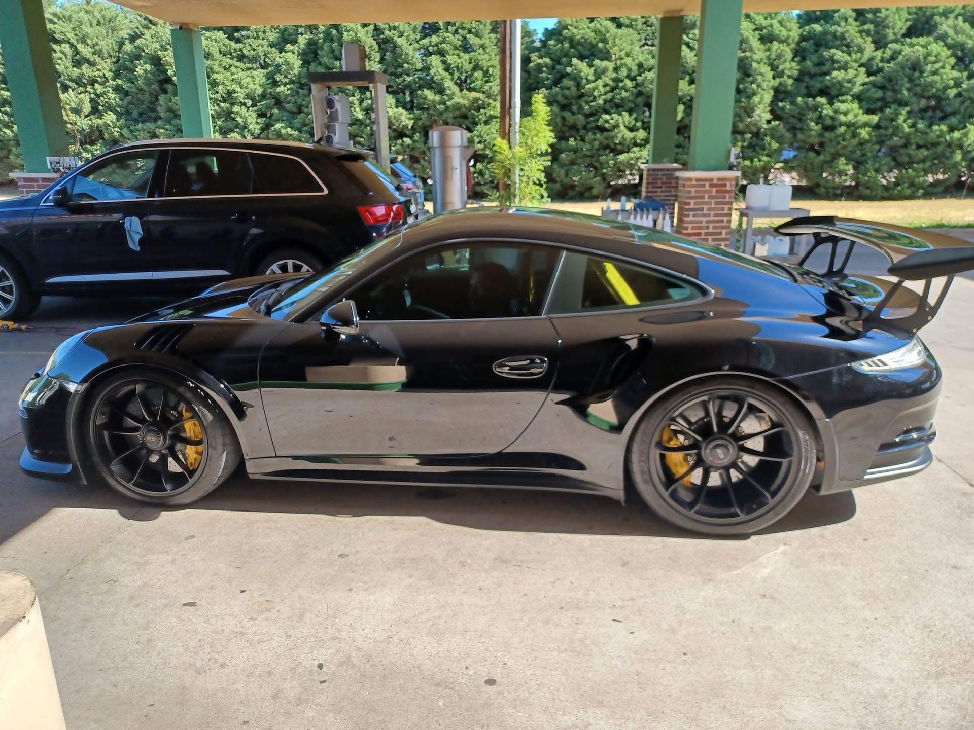 Black Porsche sports car parked near a building with a large spoiler and yellow accents. A black SUV is visible in the background.