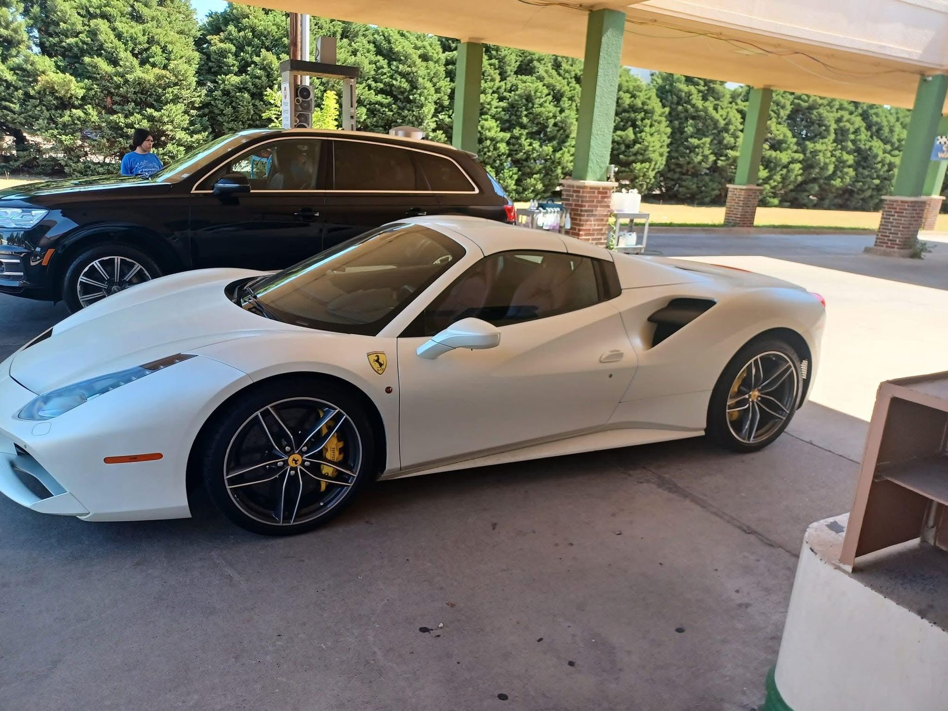 White Ferrari parked under a canopy; a black SUV is behind it. The Ferrari has yellow brake calipers.