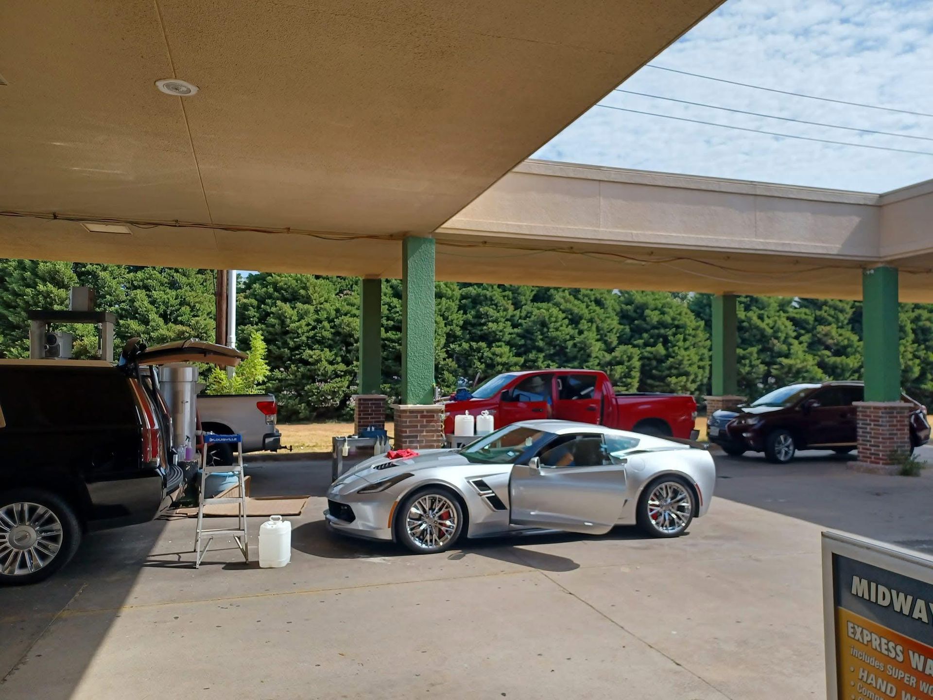 Silver sports car at a car wash, with other vehicles in the queue. Green pillars support the roof; trees in the background.