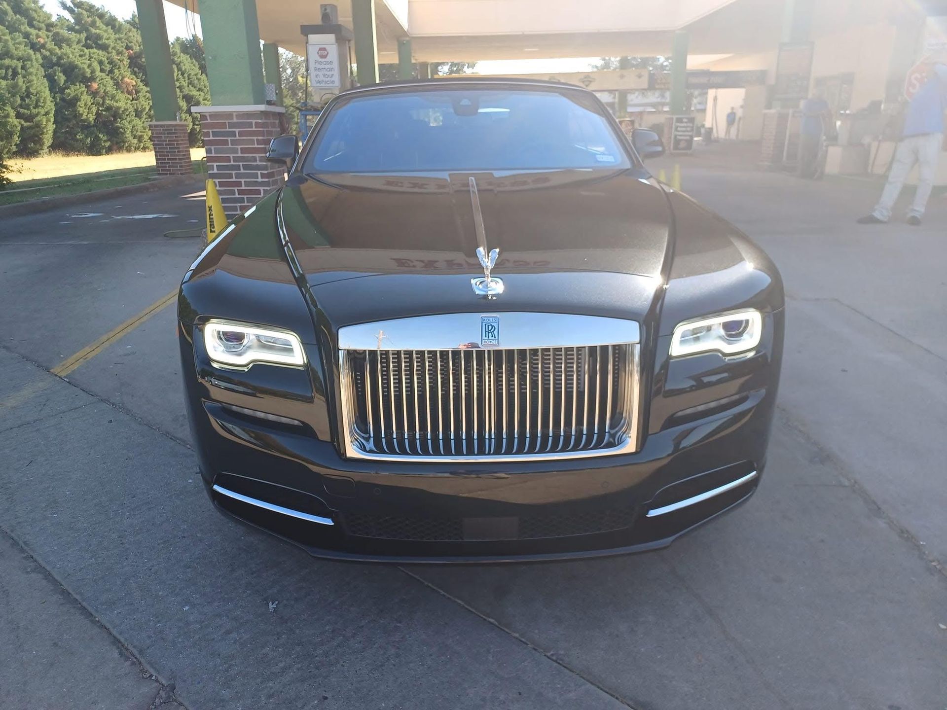 Black Rolls-Royce car parked at a gas station, viewed from the front, with the signature silver grille and hood ornament.