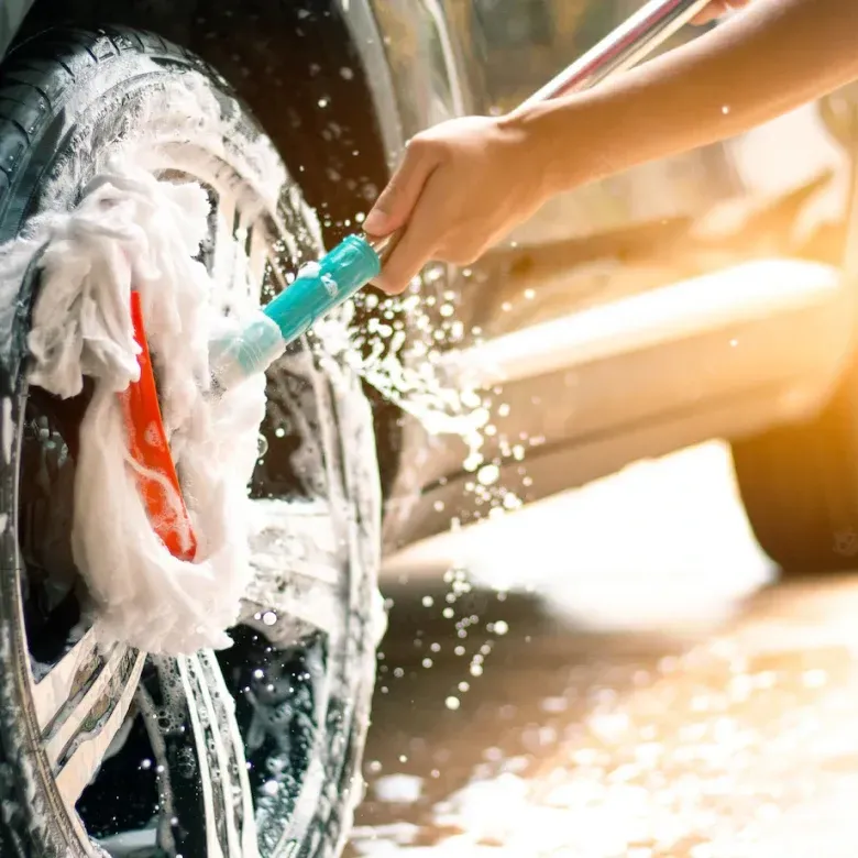 A person washing a car wheel with a soapy brush outdoors, water spraying.