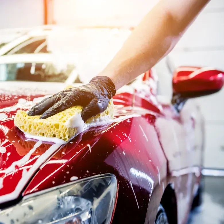 Hand in black glove washes a red car with a soapy sponge.