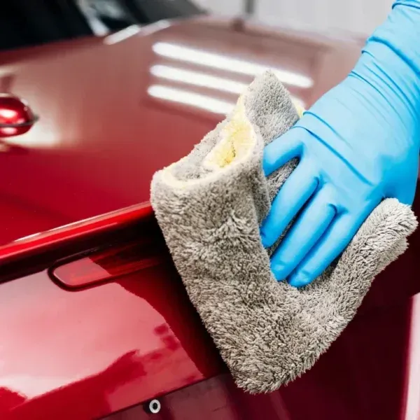 Person wearing blue gloves wiping a shiny red car with a gray microfiber cloth.