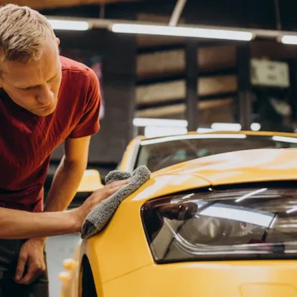 Man in red shirt wiping down a yellow car with a gray cloth in a garage.