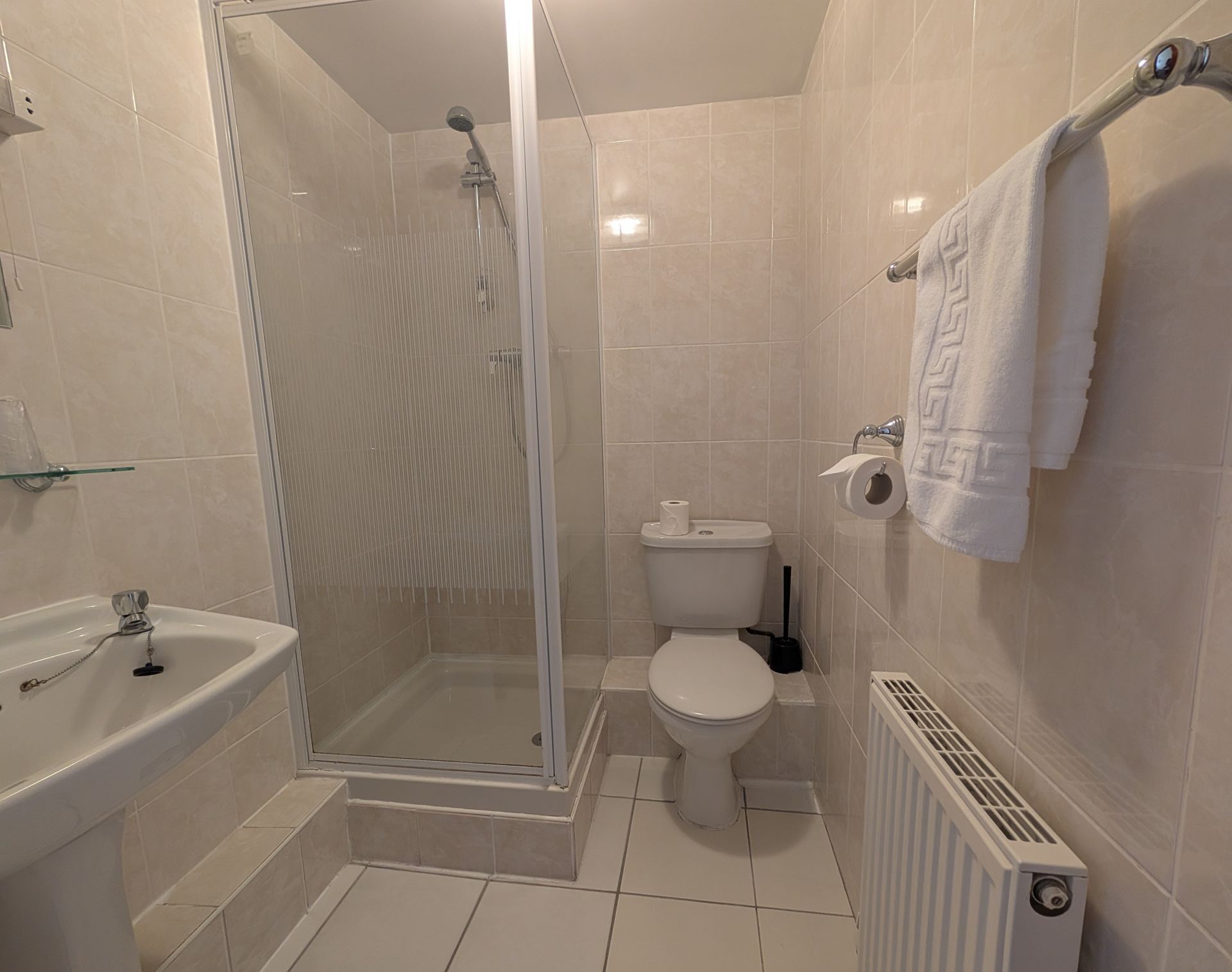 Small bathroom with a shower, toilet, sink, and towel rack. Off-white tiles and walls, white fixtures, and a radiator.