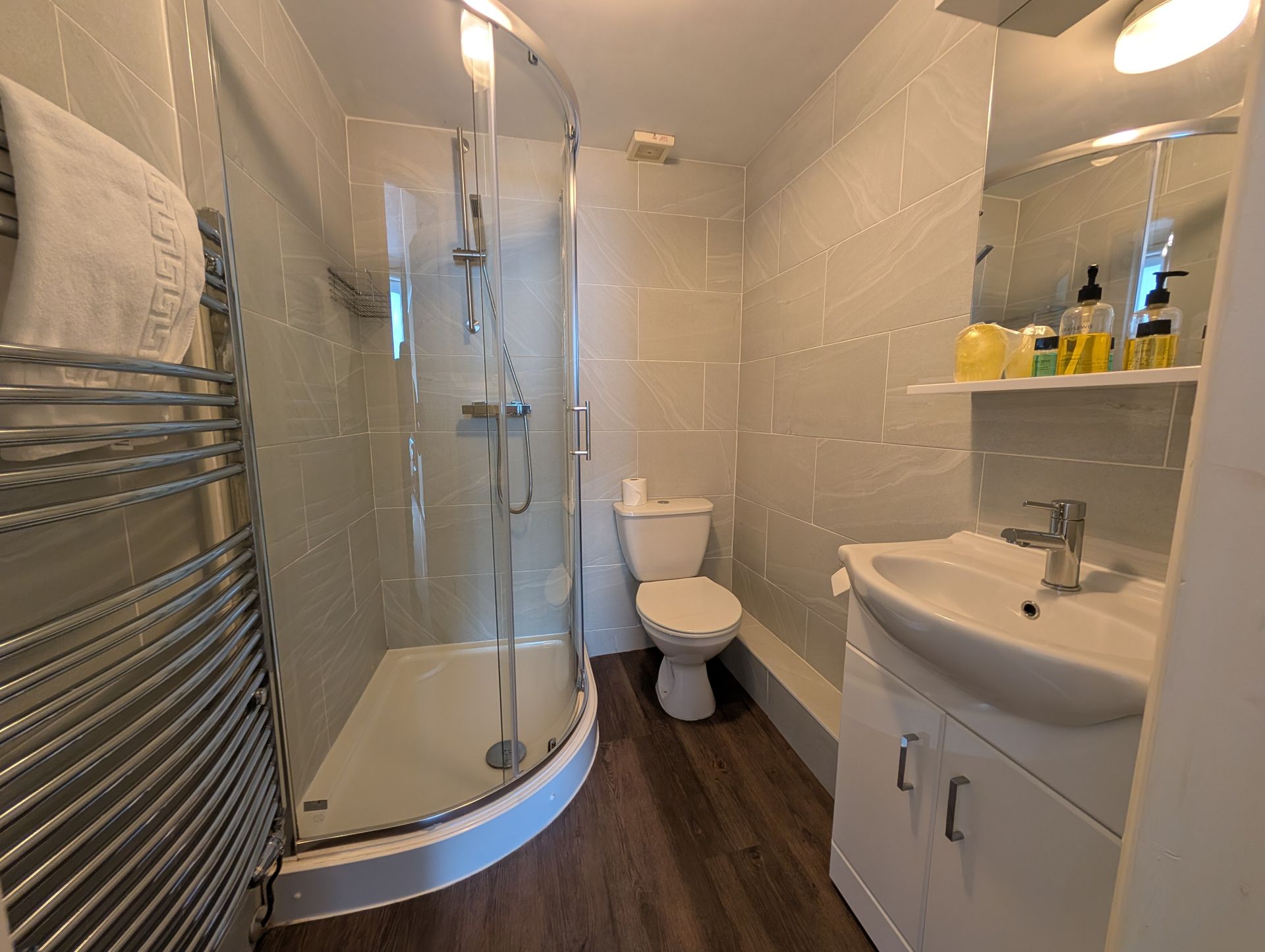 Small bathroom with shower, toilet, and sink. Chrome towel rack, white and gray walls, dark wood-look floor.