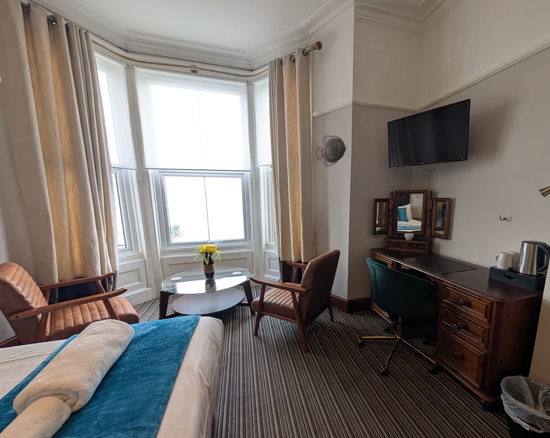 Hotel room with a bay window, table, chairs, desk, TV, and bed with blue blanket.