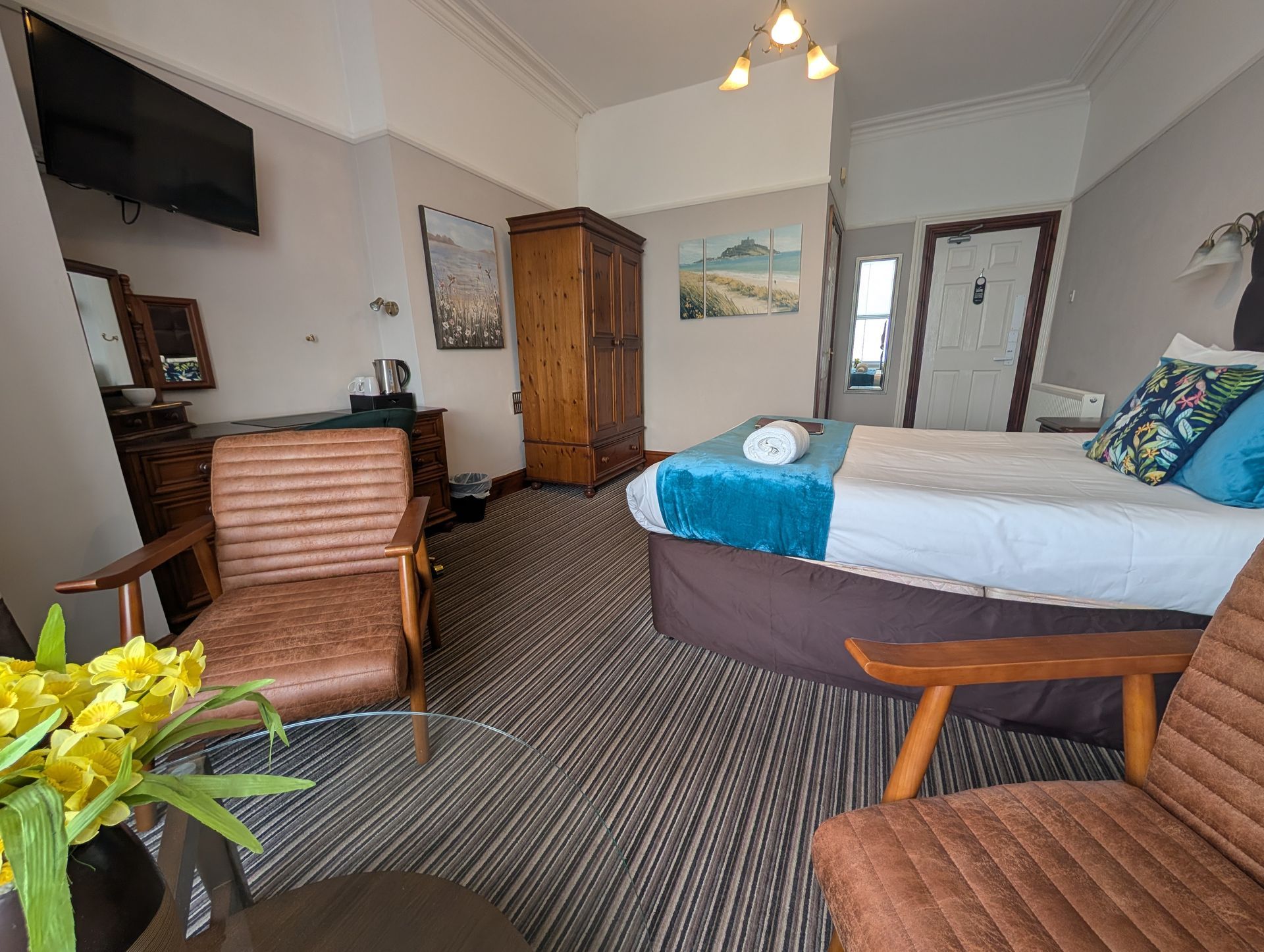 Hotel room with a bed, two chairs, a wardrobe, and a TV. The room has neutral colors with teal and brown accents.