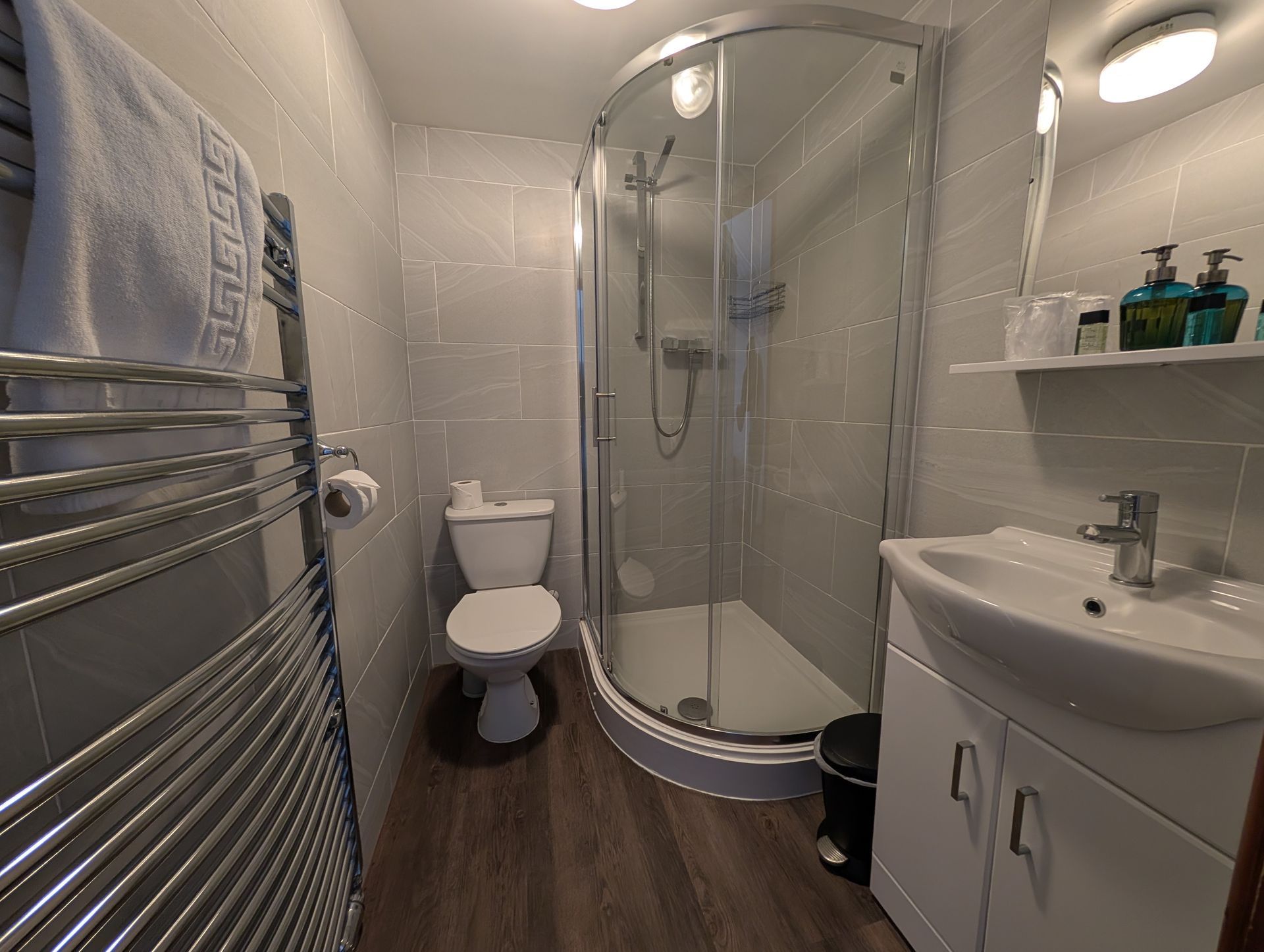 Small bathroom with shower, toilet, sink, and towel warmer; light gray tiles and wood-look flooring.