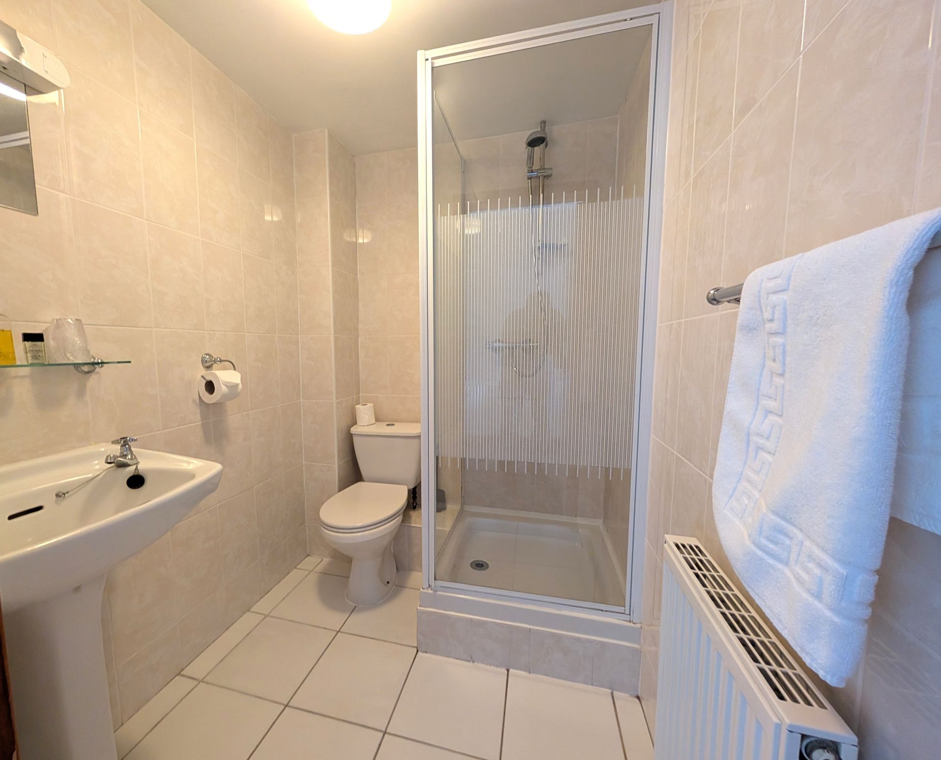 Small, tiled bathroom with a sink, toilet, shower, and towel on a radiator.