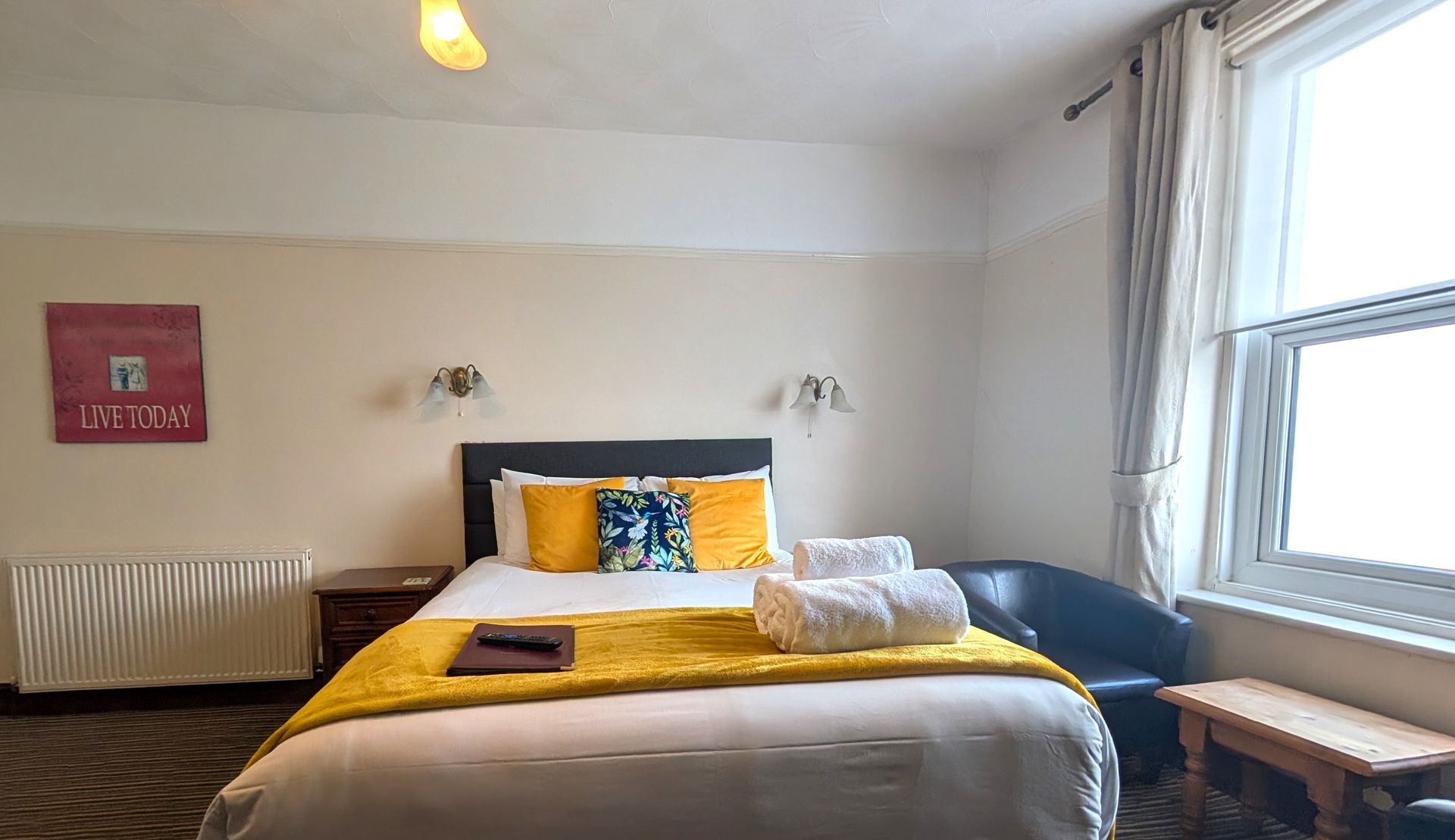 Bedroom with double bed, yellow and blue accents, white towels, a window, and a small sitting area.