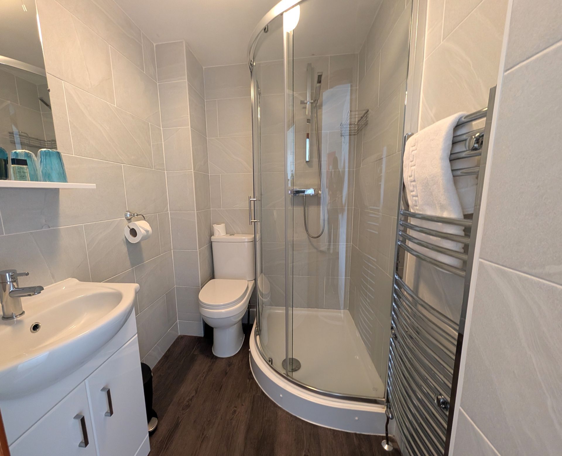 Small bathroom with a toilet, shower, sink, and towel rack. Gray and white color scheme, dark wood-look floor.