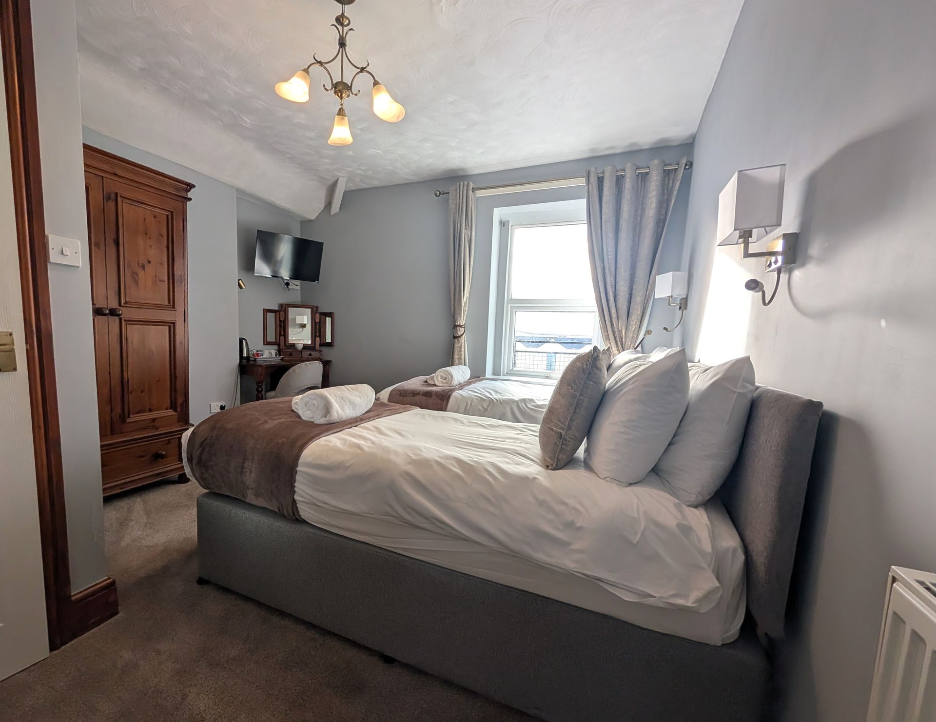 Bedroom with two beds, gray walls, wooden wardrobe, window with curtains, and small television.